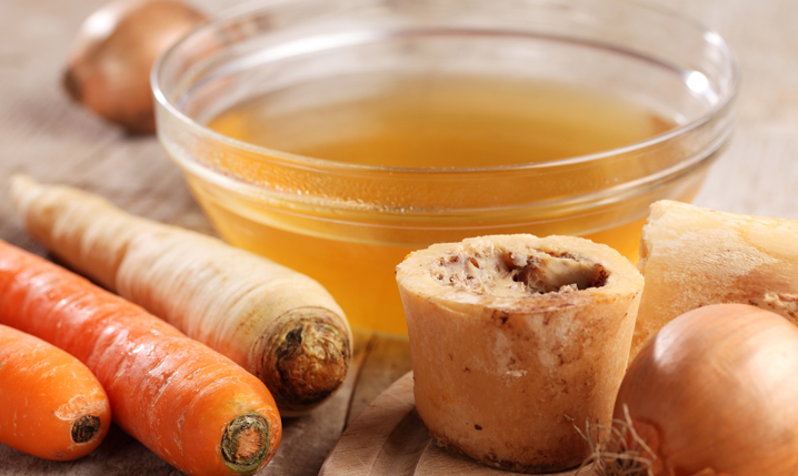 a bowl of beef broth next to common ingredients such as carrots, onions, and beef bone containing marrow