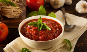marinara sauce in a bowl with common ingredients in the background such as tomatoes and garlic